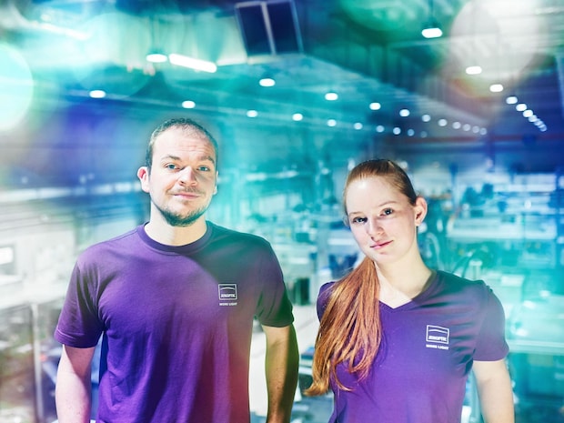 2 Jenoptik-Mitarbeiter im violett-farbenen Shirt schauen in die Kamera, Produktionshalle im Hintergrund